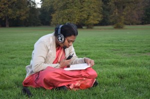 லண்டன் புள்வெளியில் அமர்ந்து காதில் ஹெட்போன் செருகியபடி ஒலிப்பதிவு செய்யும் திருமதி. உஷா ராமகிருஷ்ணன் அவர்கள்